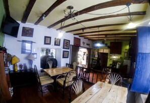 Un coin repas rustique avec des tables et des chaises en bois, un lustre et une cuisine visible au fond.