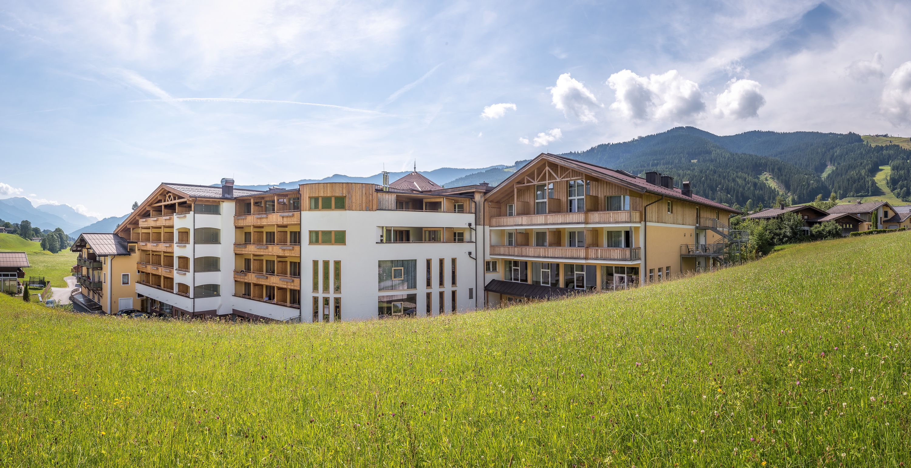 A modern building with multiple balconies and a flat roof, surrounded by a lush green field and mountains in the background.