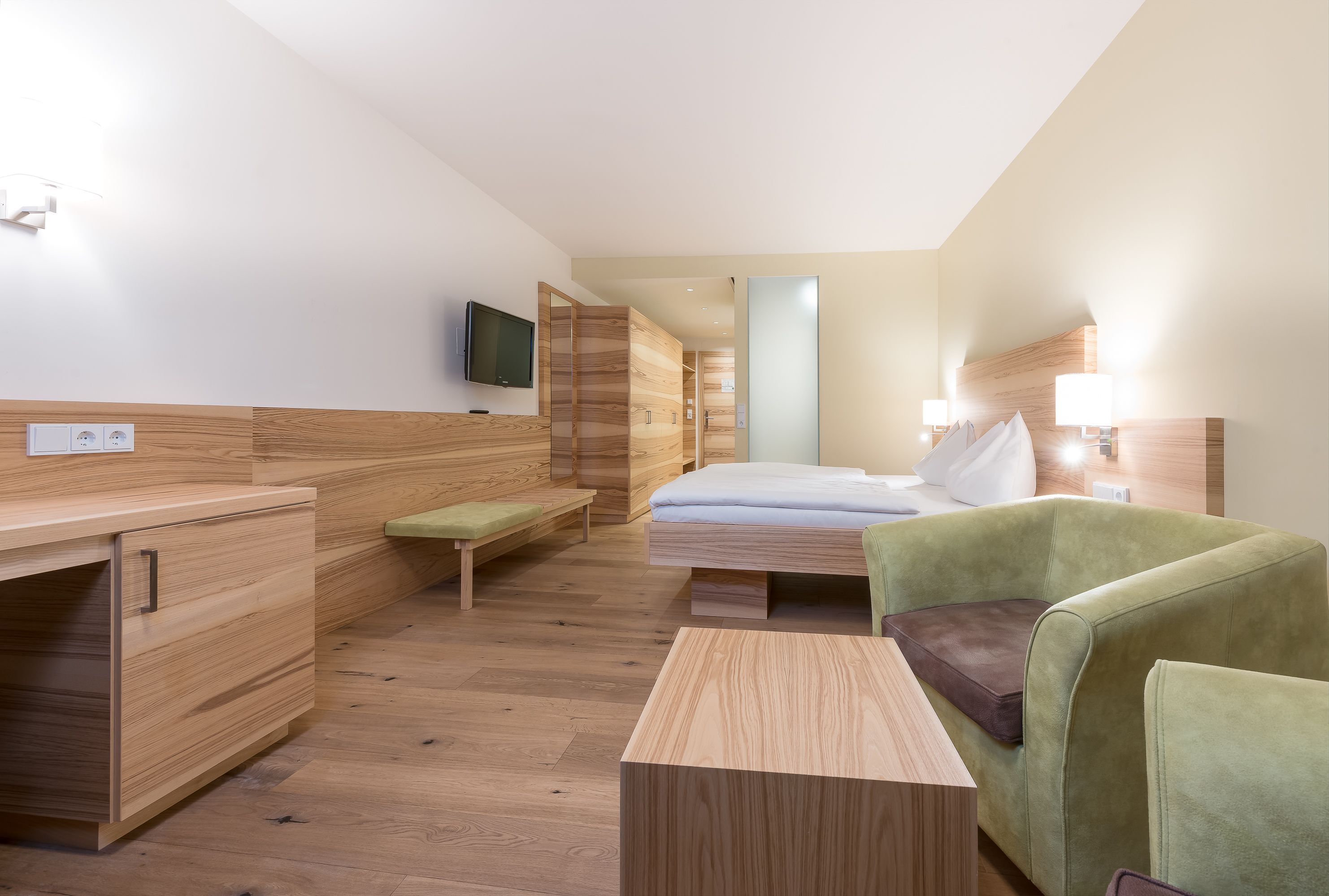 A modern hotel room with a wooden floor, a bed with a green and brown upholstered headboard, a desk with a flat-screen TV, and a bench.