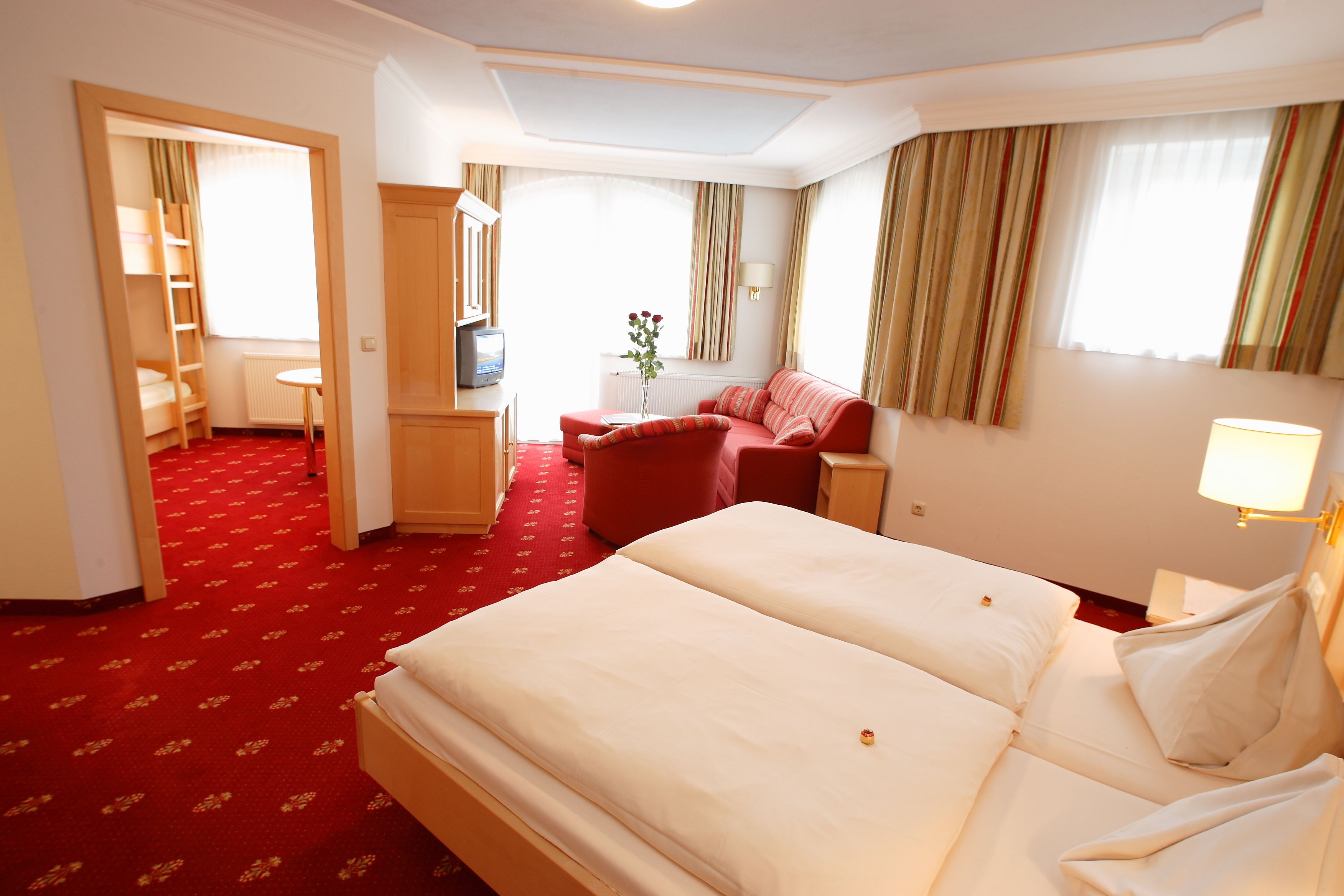 A hotel room with a large bed, a desk, a chair, and a small table. There is a window with curtains and a radiator.