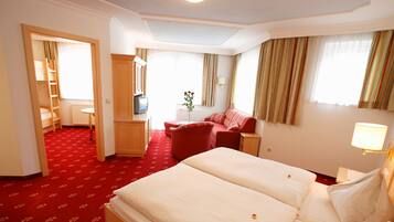 A hotel room with a large bed, a desk, a chair, and a small table. There is a window with curtains and a radiator.