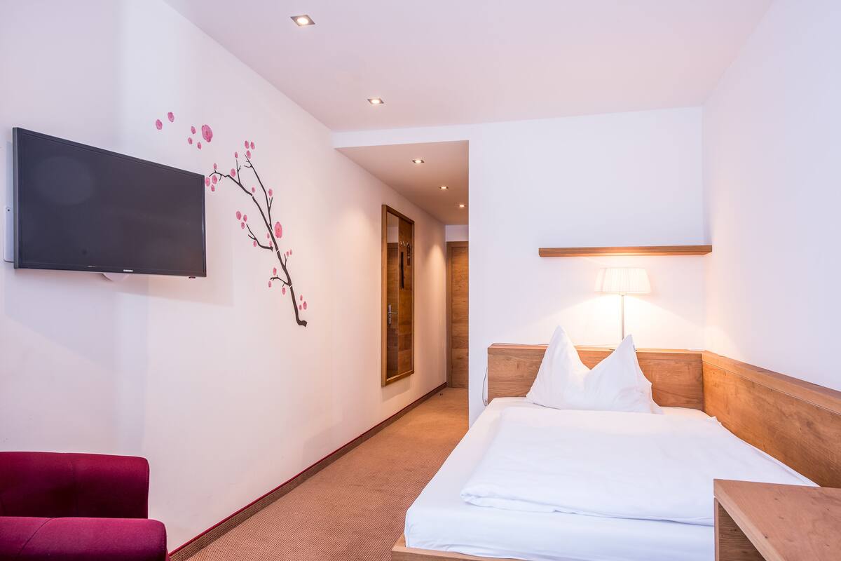 A hotel room with a bed, a TV mounted on the wall, a lamp, and a red chair.