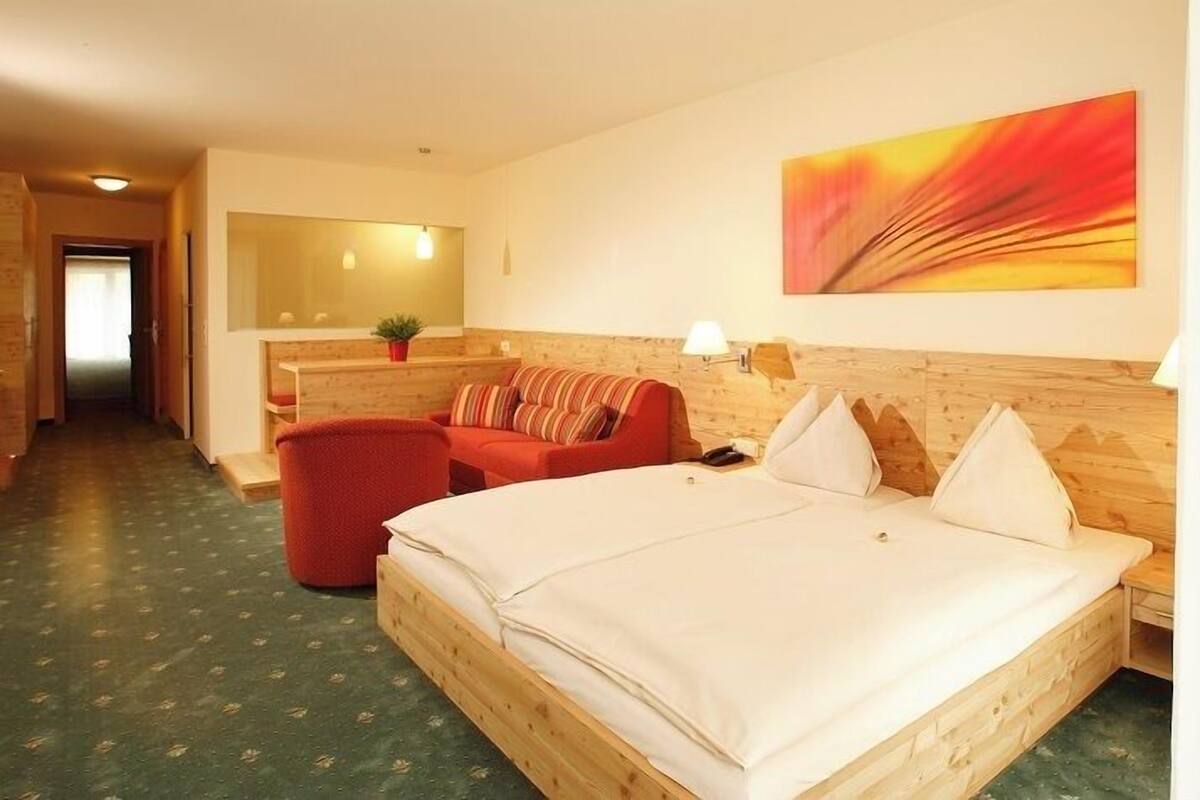 A hotel room with a wooden headboard, a large bed, a red armchair, and a painting on the wall.