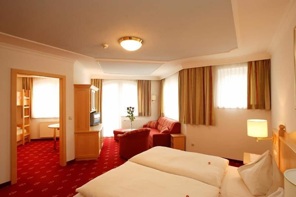 A hotel room with two beds, a red carpet, a desk with a lamp, a TV, and a seating area with a red sofa and armchairs.