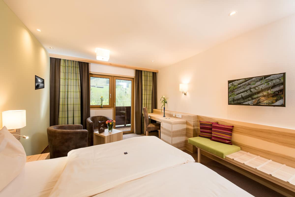 A hotel room with a large bed, a desk, a chair, a small table with a vase, and a bench with pillows.