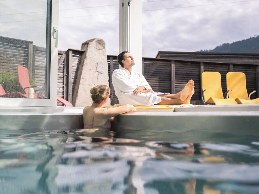 Zona de piscina con una persona descansando en una silla lounge, otra persona en el agua y una montaña al fondo.