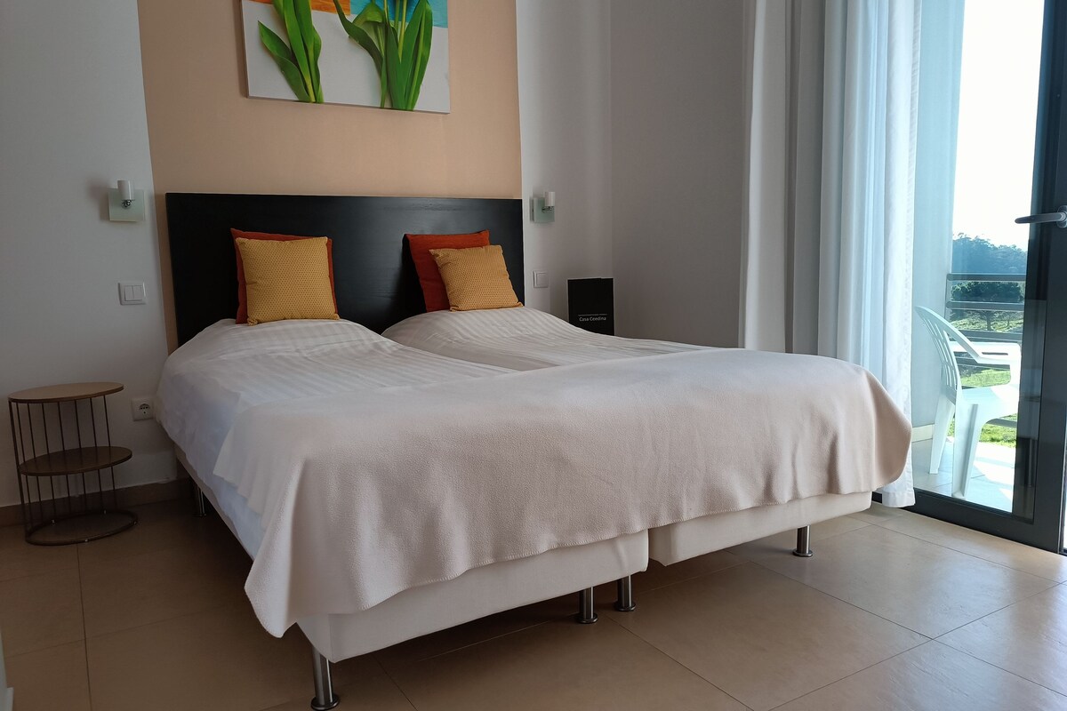 Ein Doppelbett mit weißen Leinen, ein schwarzes Kopfteil und zwei dekorative Kissen. Neben dem Bett steht ein kleiner runder Tisch, und an der Wand hängt ein Blumenbild.