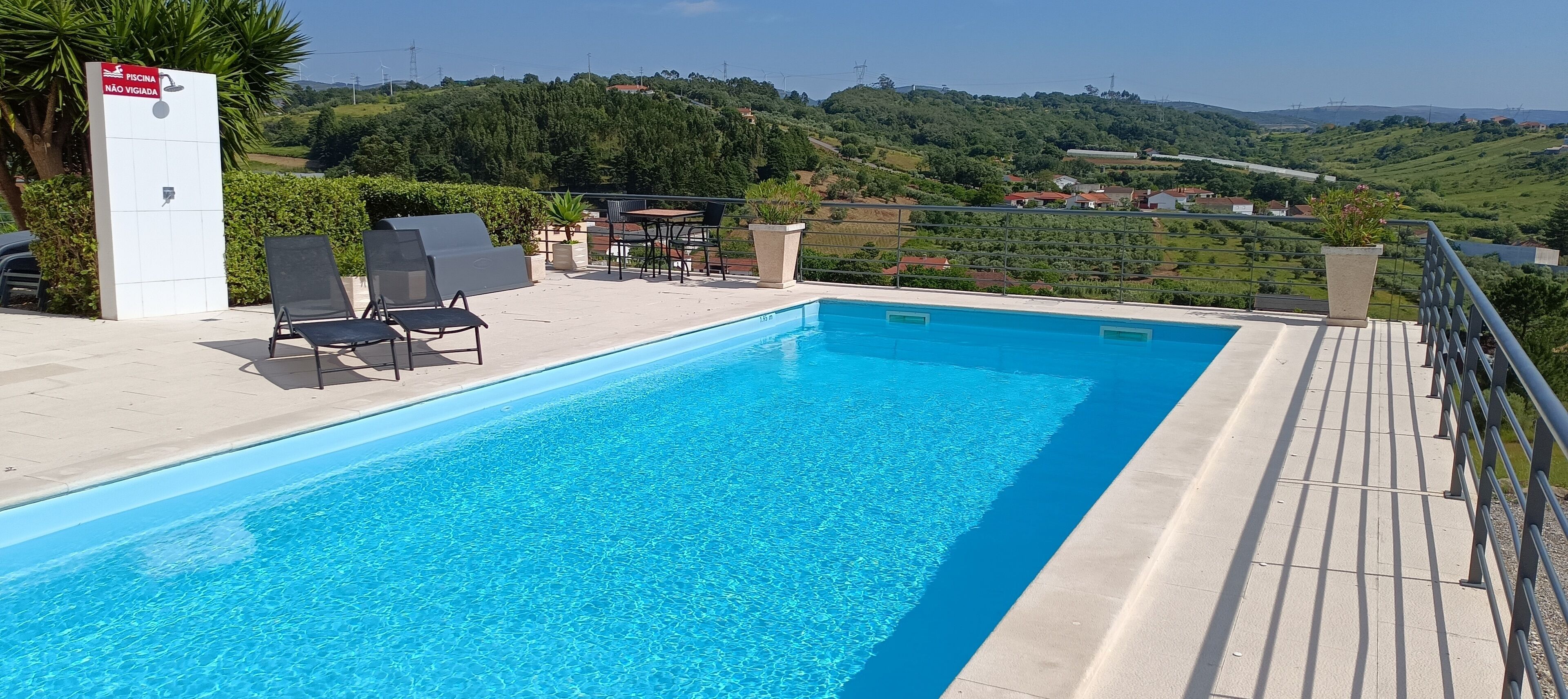 A swimming pool with lounge chairs and a table, surrounded by a railing, with a scenic view of hills and houses in the background.