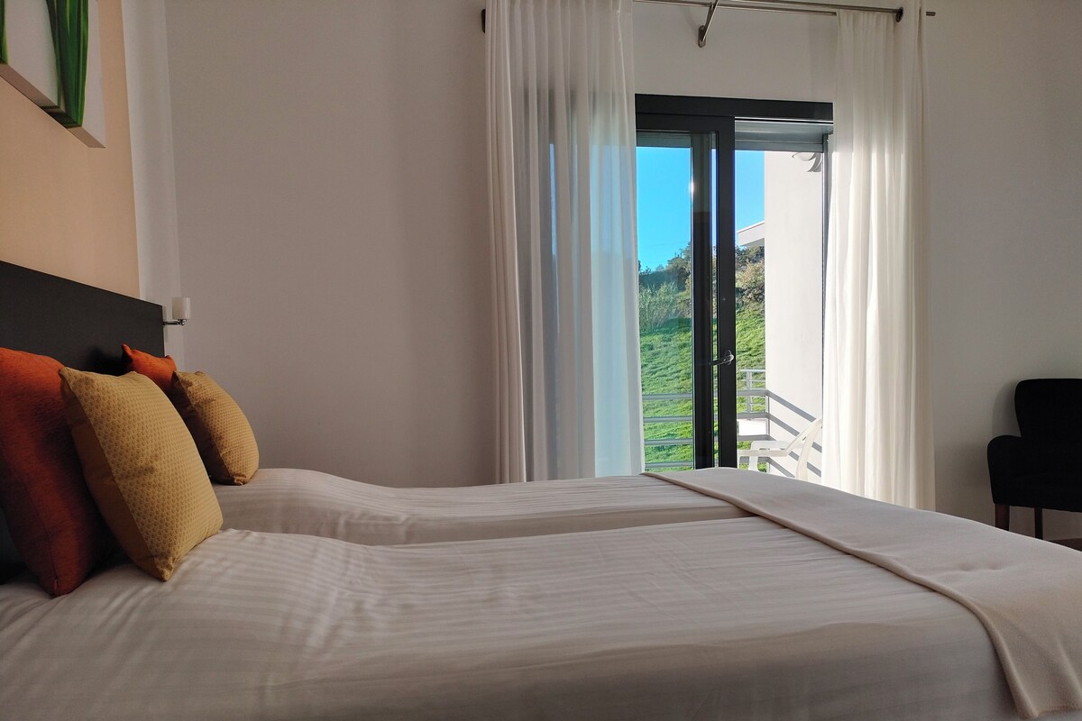 A bedroom with two beds, a view of a balcony, and a chair.