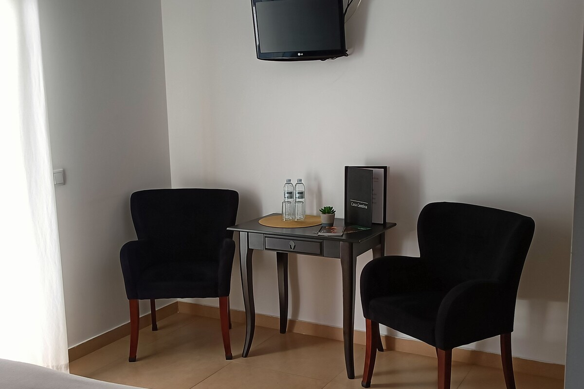 A hotel room with two black armchairs, a small table with a water bottle, a plant, and a framed picture.