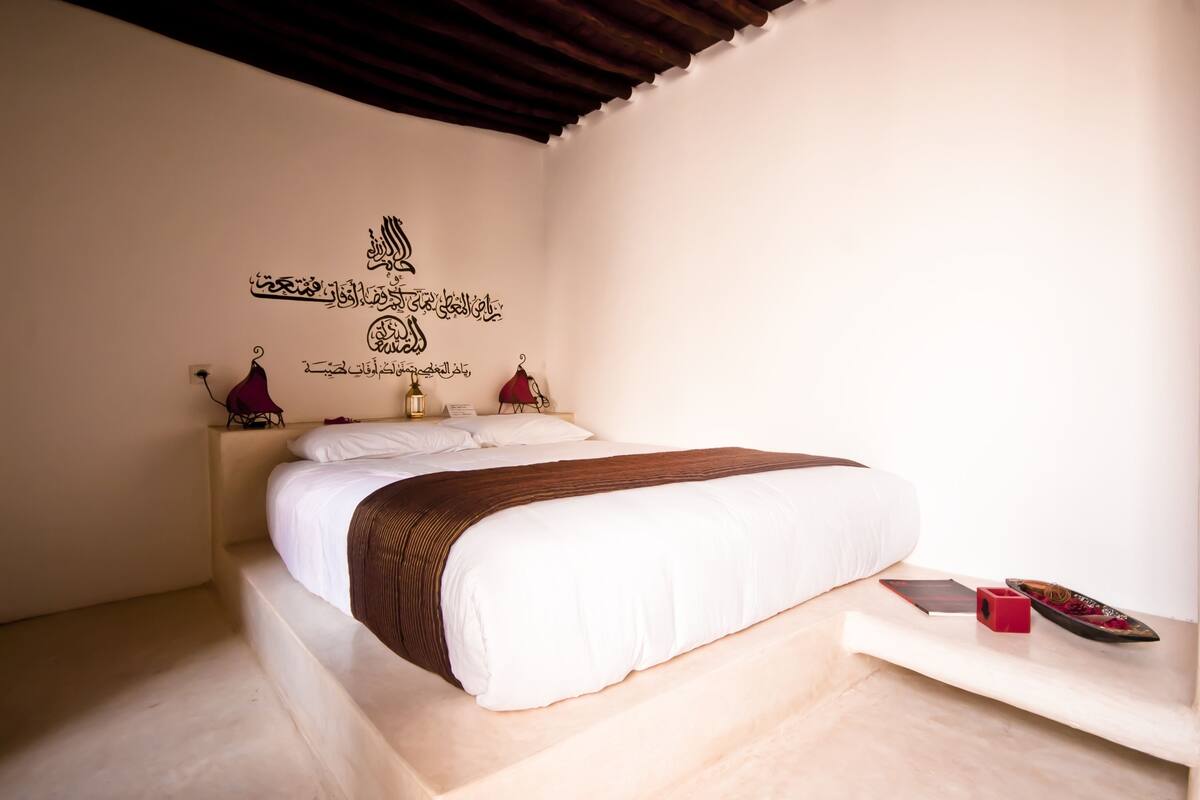 A neatly made bed with a brown headboard, a bedside table with a lamp, and a wall with Arabic calligraphy.