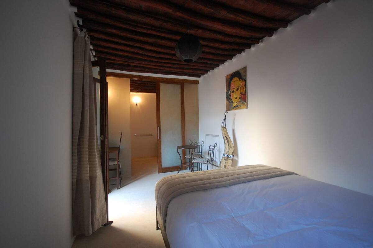 A bedroom with a bed, a desk, and a chair, a painting on the wall, and a wooden ceiling.