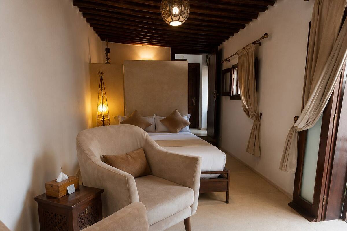 A bedroom with a bed, two armchairs, a bedside table with a tissue box, a hanging light fixture, and a window with curtains.