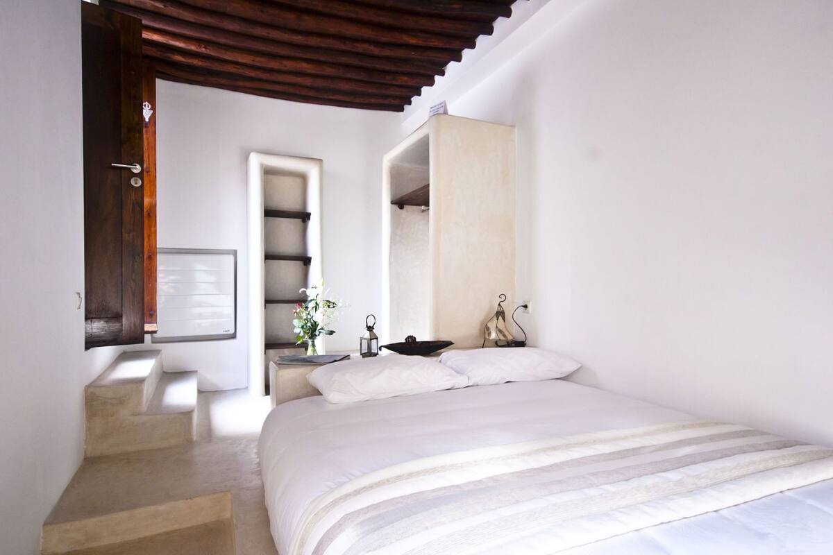 A bedroom with a bed, a nightstand, a vase with flowers, a lamp, and a wooden door.