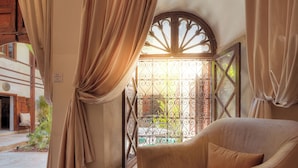 Una habitació amb vista per una finestra amb reixes, una cadira amb coixí i una taula petita.