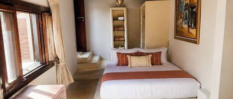 A bedroom with a bed, a window, a wardrobe, a painting, and a hanging light fixture.