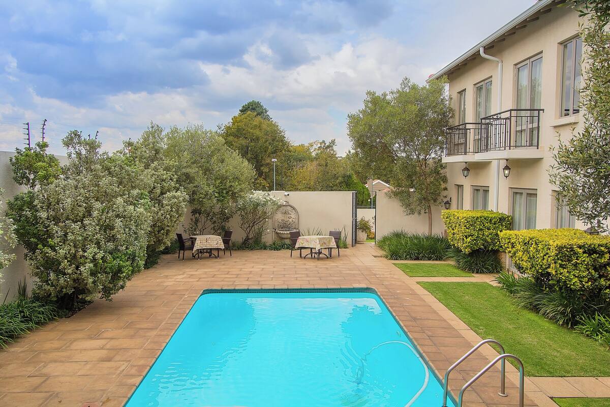 A swimming pool with a tiled deck, surrounded by greenery and a building with a balcony.