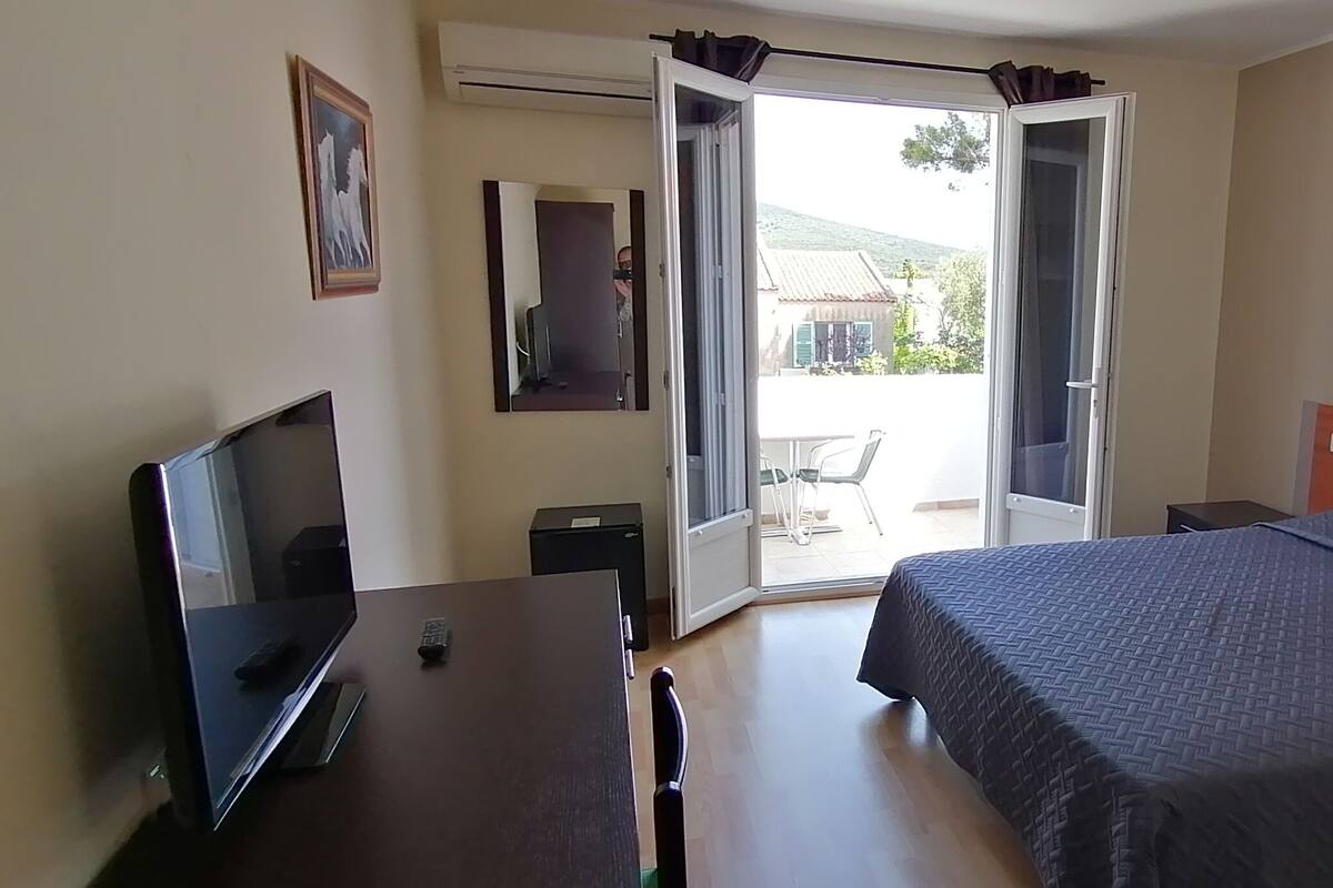 A hotel room with a bed, a desk with a TV, a chair, and a door leading to a balcony.