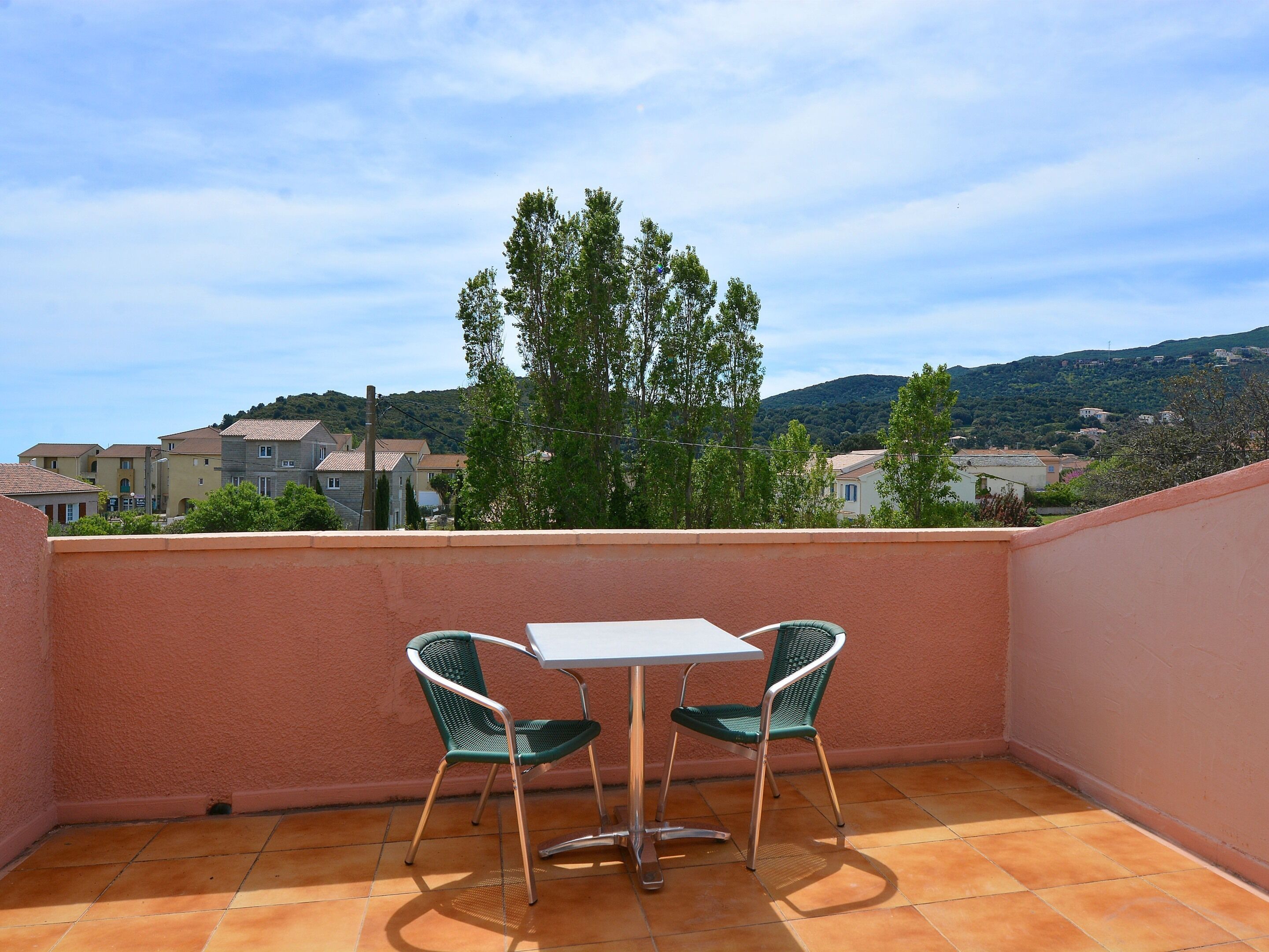 A balcony with a table and two chairs, overlooking a residential area with houses and trees.