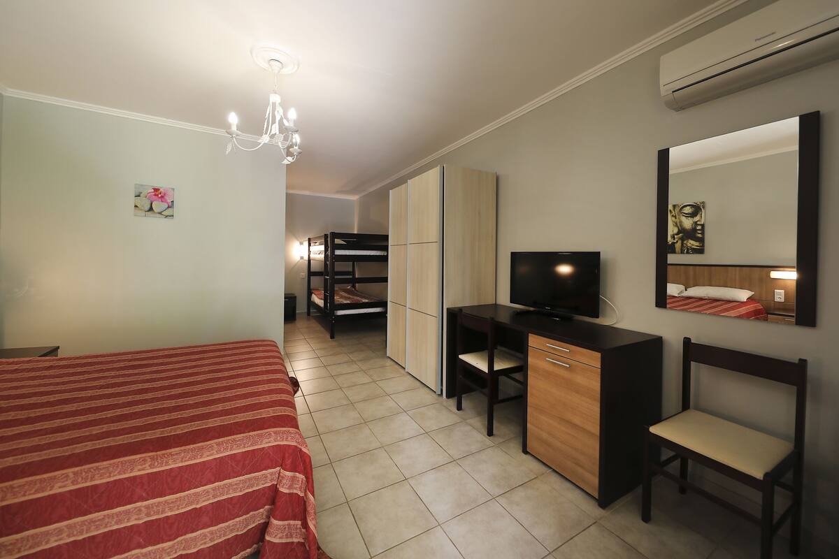 A hotel room with a bed, desk, chair, TV, wardrobe, and an air conditioning unit.