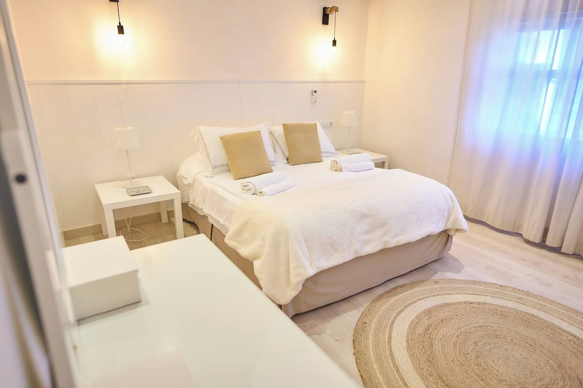 A hotel room with a bed, white nightstands, a bedside table, and a large window with curtains.