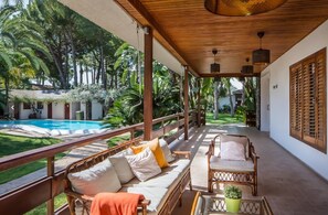 A covered outdoor seating area with wicker furniture, a pool, and lush greenery in the background. - El Oasis (L'Eliana)