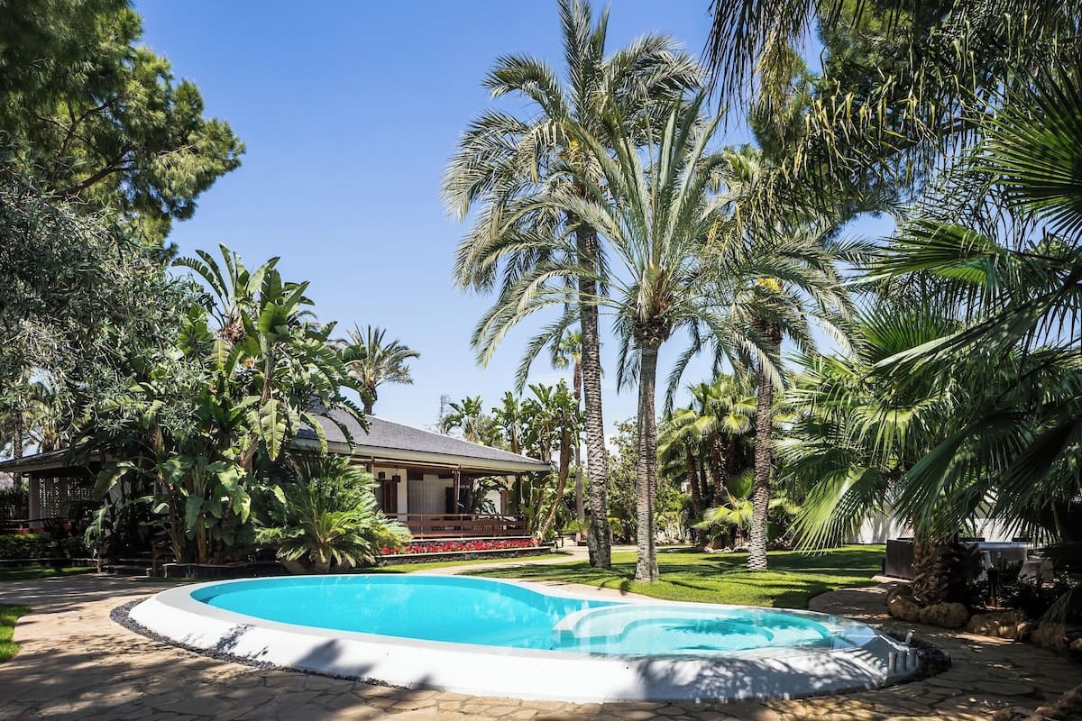 A pool surrounded by palm trees and a house with a covered deck.