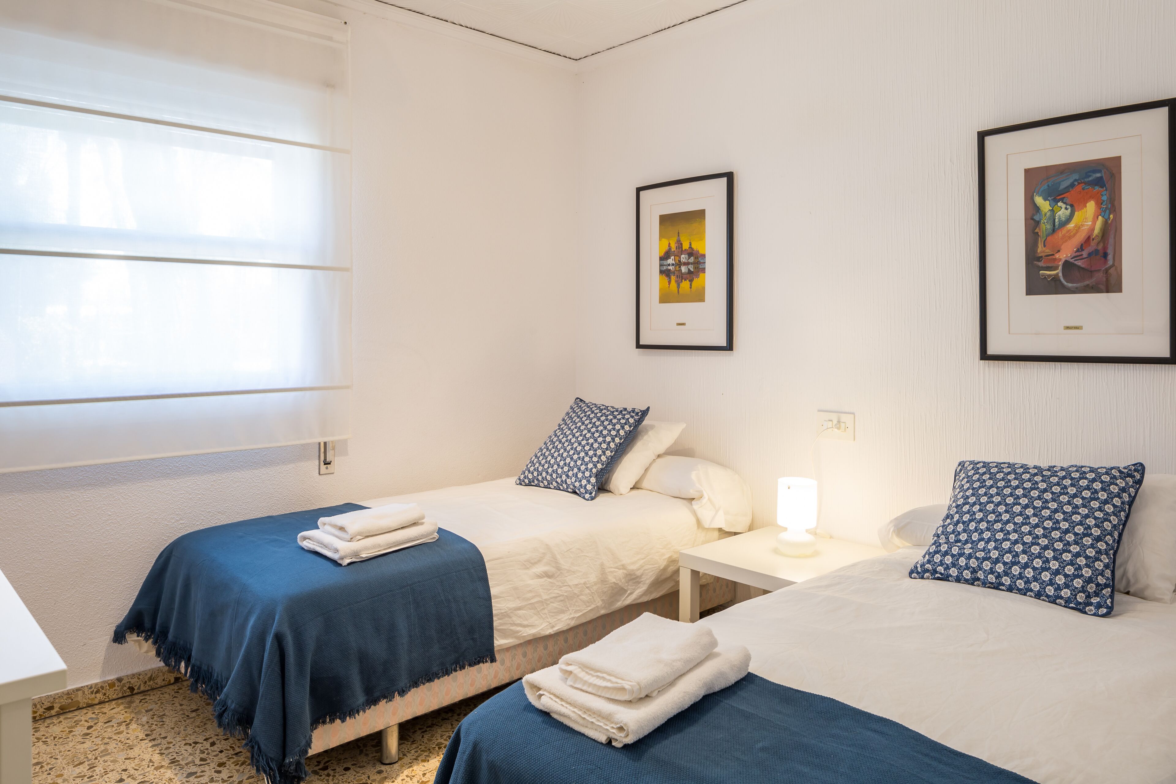 Une chambre d’hôtel avec deux lits, une fenêtre avec des stores, une table de chevet avec une lampe et des œuvres d’art encadrées accrochées aux murs.