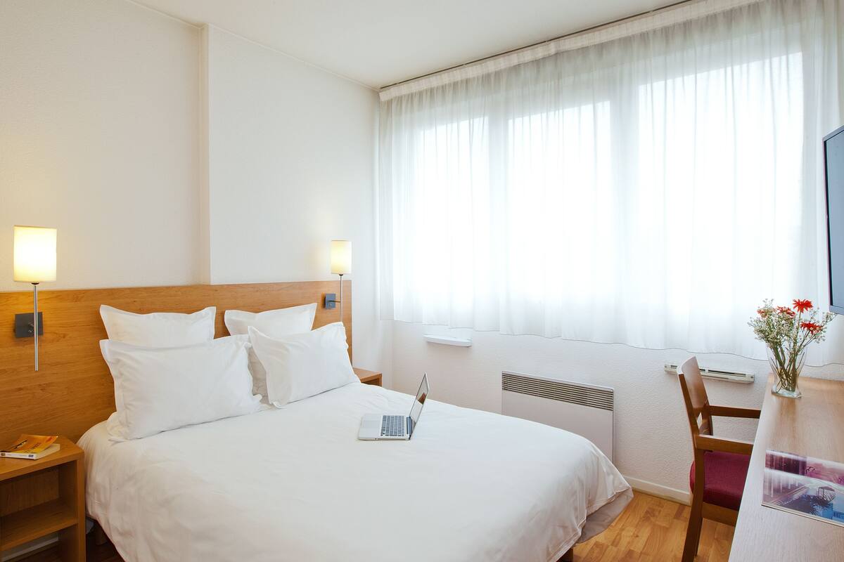Ein Hotelzimmer mit einem großen Bett, einem Schreibtisch mit einem Laptop, einem Stuhl und einem Fenster mit transparenten Vorhängen.