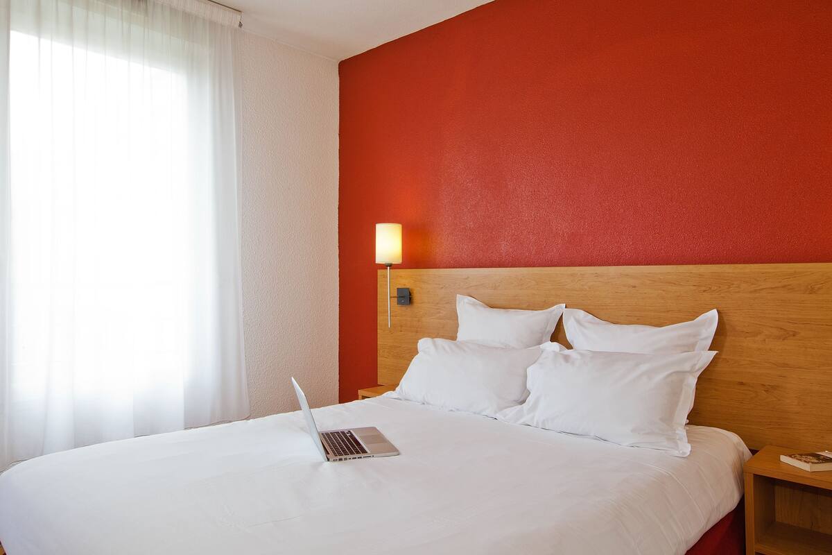 Ein Hotelzimmer mit einem Bett, einem Laptop auf dem Bett, einem Nachttisch und einer Wandlampe.