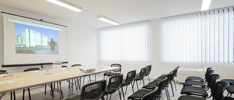 A conference room with a long table, black chairs, a projector screen, and large windows with blinds.