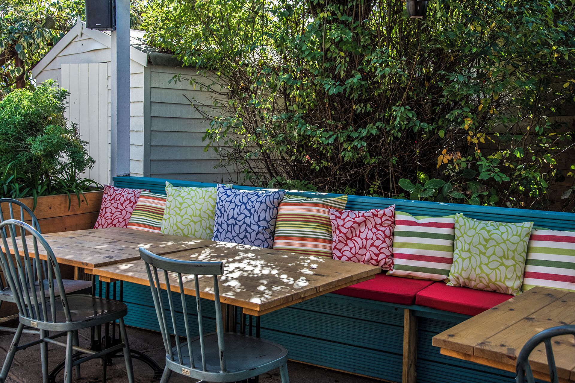 Een terras met een houten tafel en stoelen, een kleurrijke bank met patroon kussens, en een achtergrond van weelderige begroeiing.