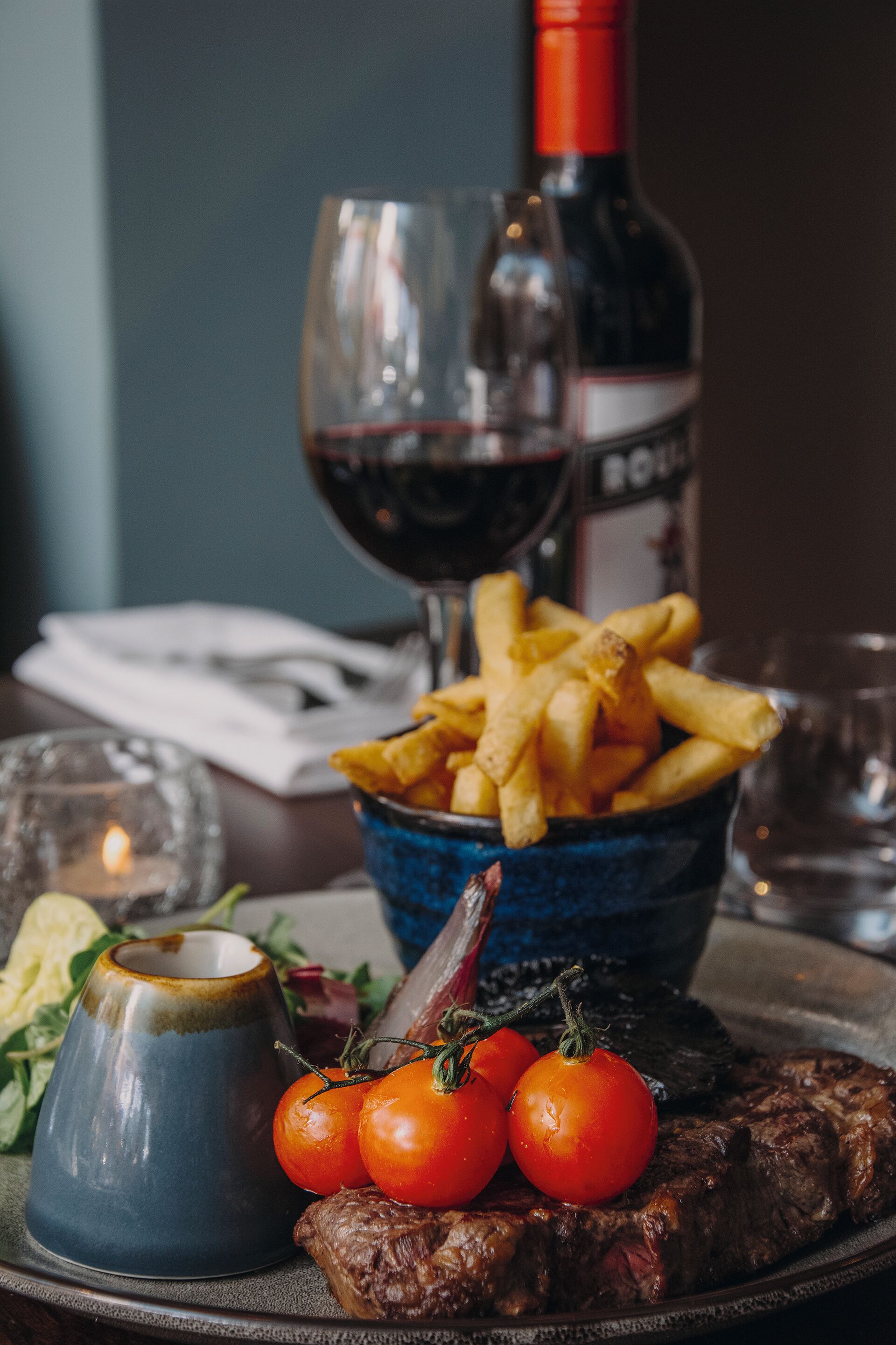 Un plato con bistec, papas fritas y tomates, una botella de vino y copas sobre una mesa.