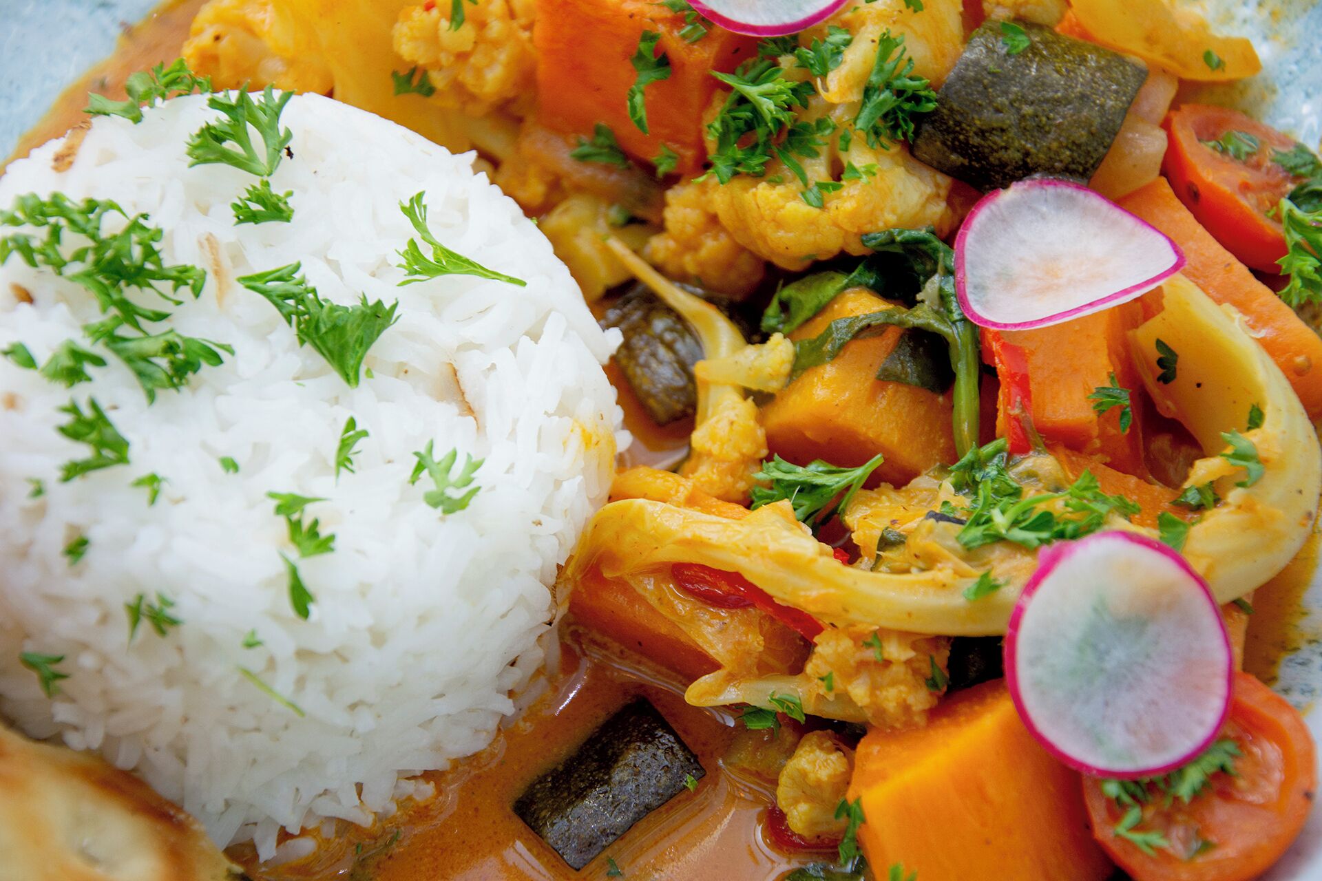 Een bord witte rijst, versierd met peterselie, geserveerd met een groentecurry met wortelen, groene bonen en een stukje vlees, mogelijk kip of tofu.