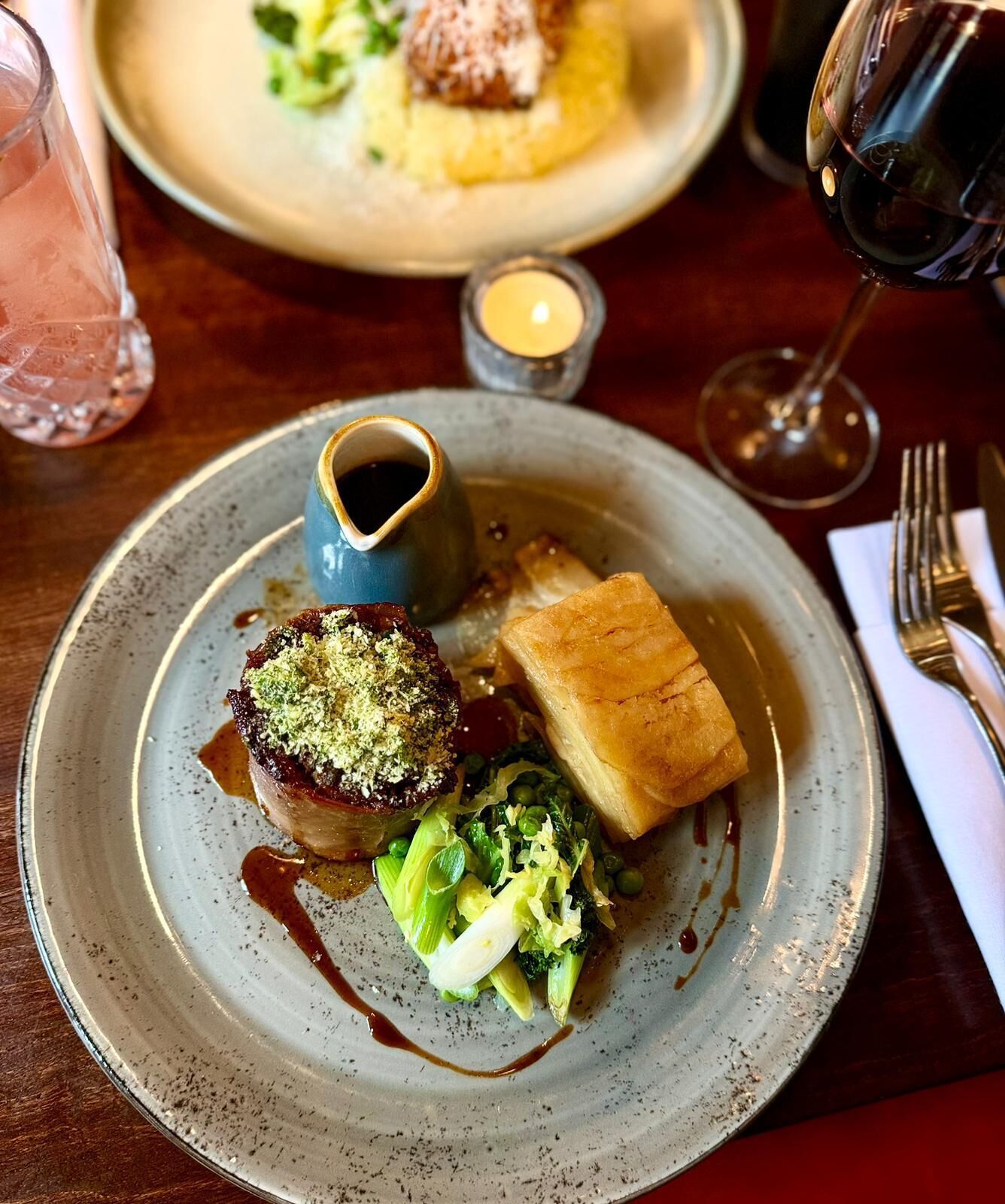 A plate of food with a piece of meat topped with green garnish, a square pastry, and a side of broccoli.