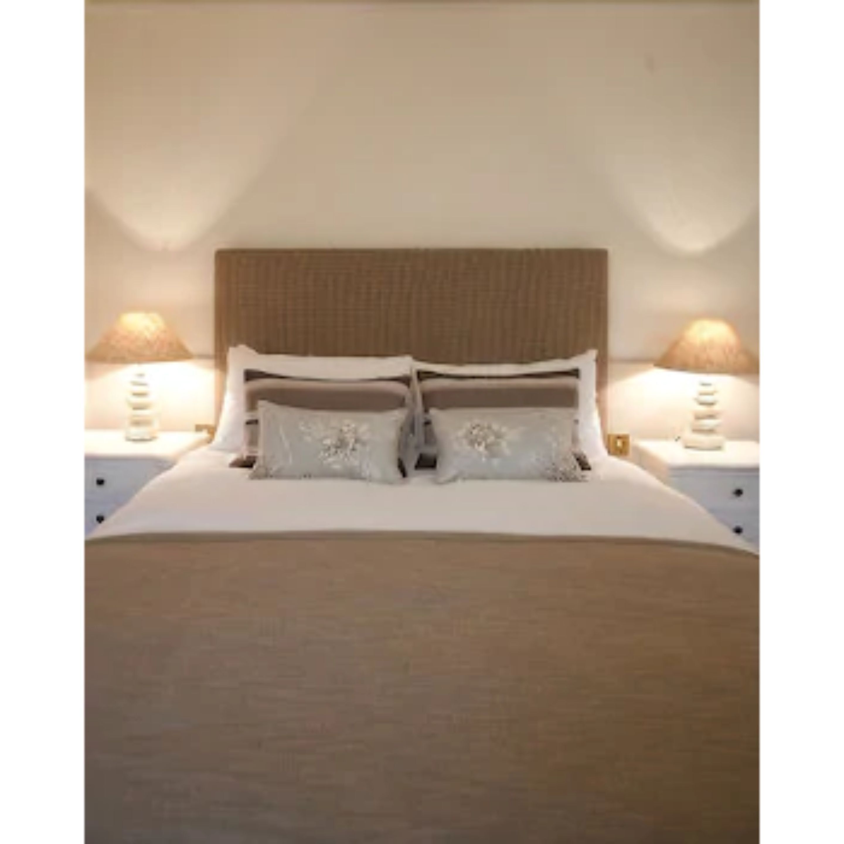 Een net opgemaakt bed met een hoofdeinde, twee nachtkastjes met lampen, en een effen muur.