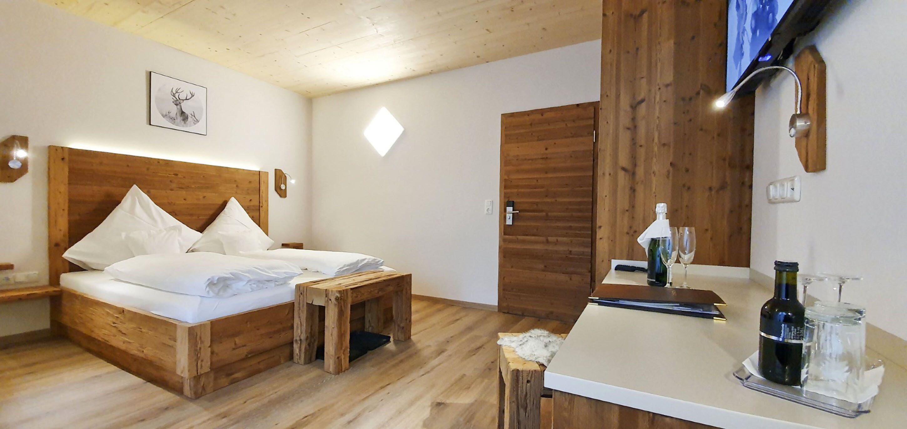 Una cama de madera con almohadas, un taburete pequeño de madera, una mesita de noche con una botella y un vaso, y un armario de madera.