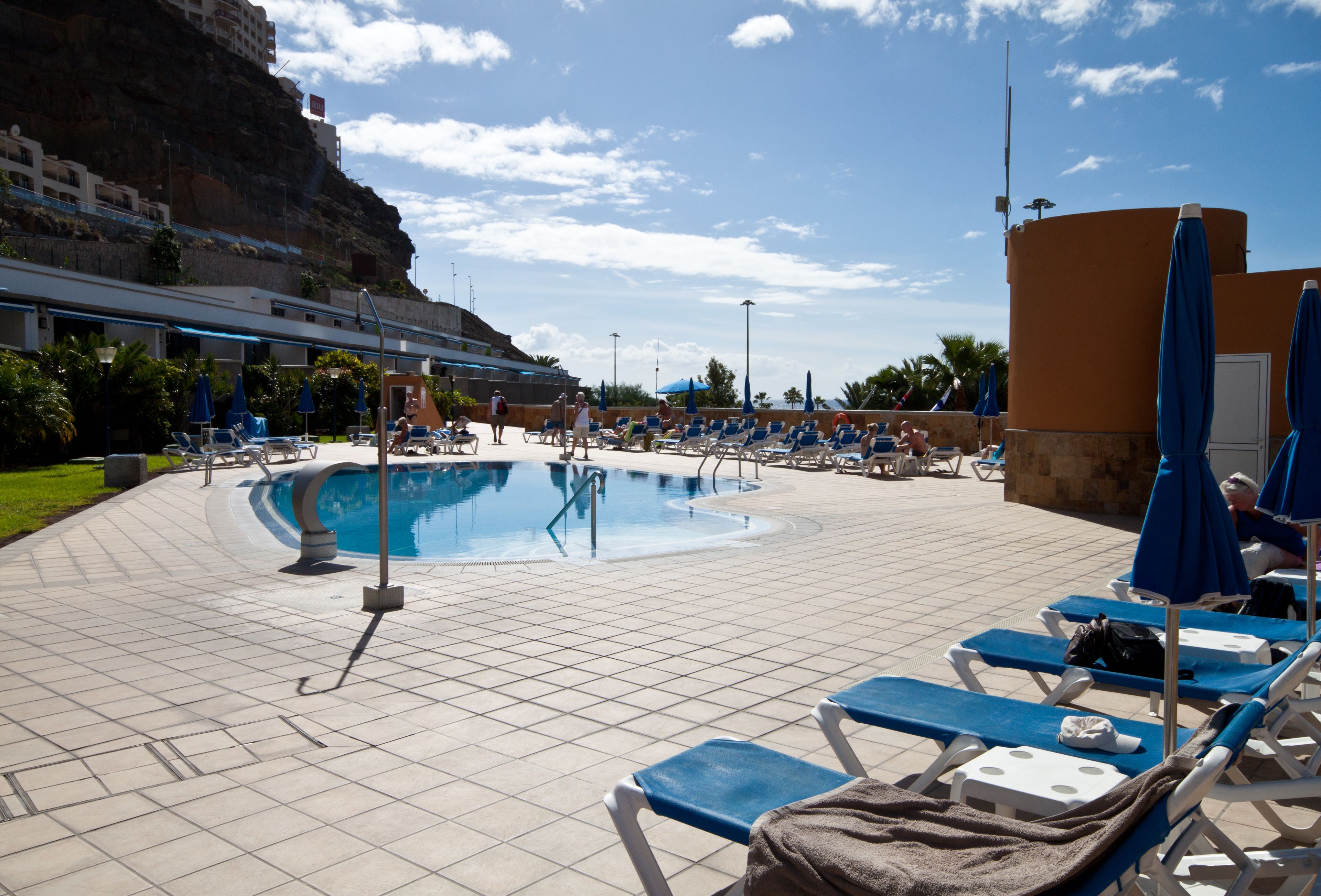 Un área de piscina con suelo de baldosas, sillas de descanso y sombrillas.
