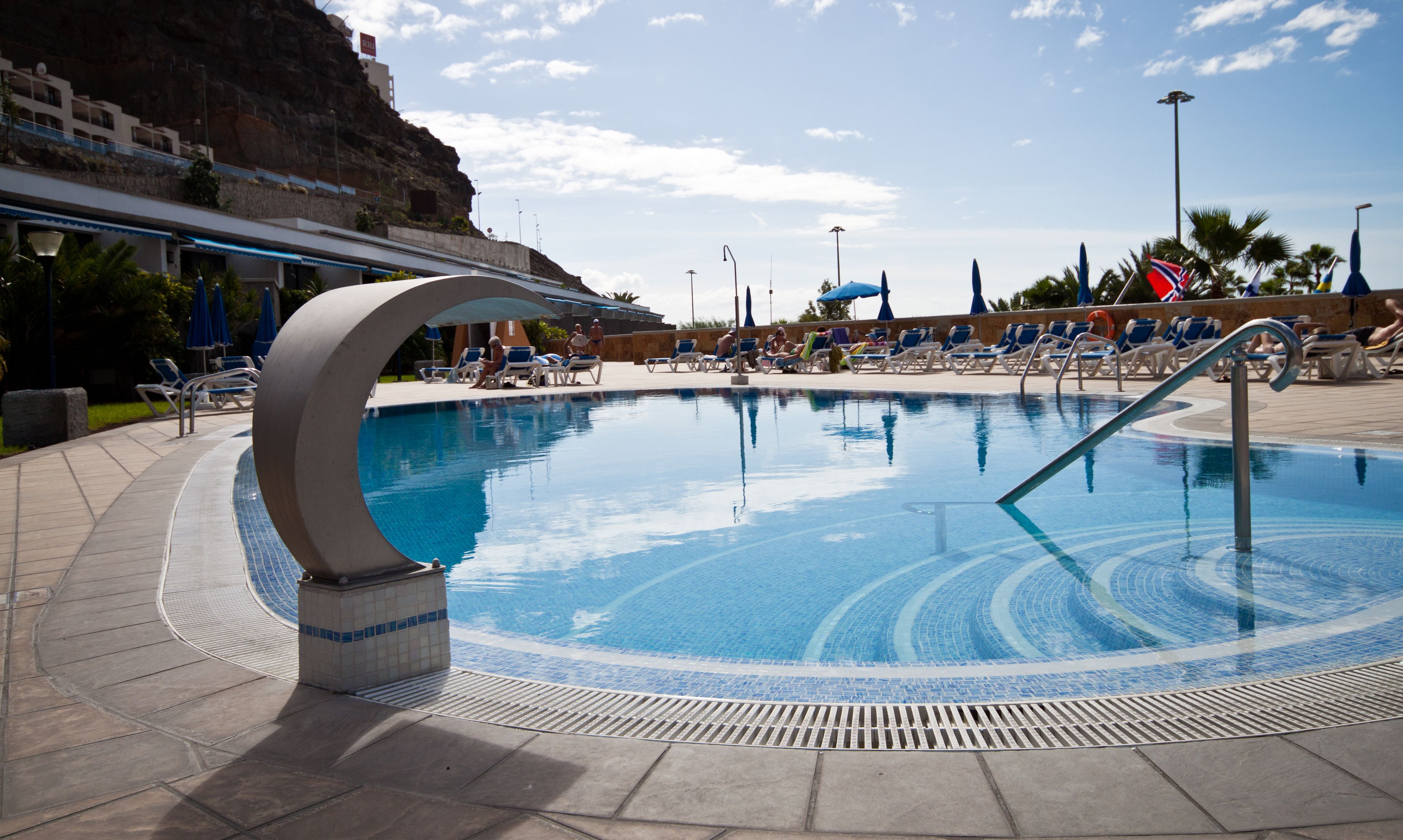 A swimming pool with a circular design, surrounded by lounge chairs and umbrellas, with a view of a rocky cliff and a clear sky.