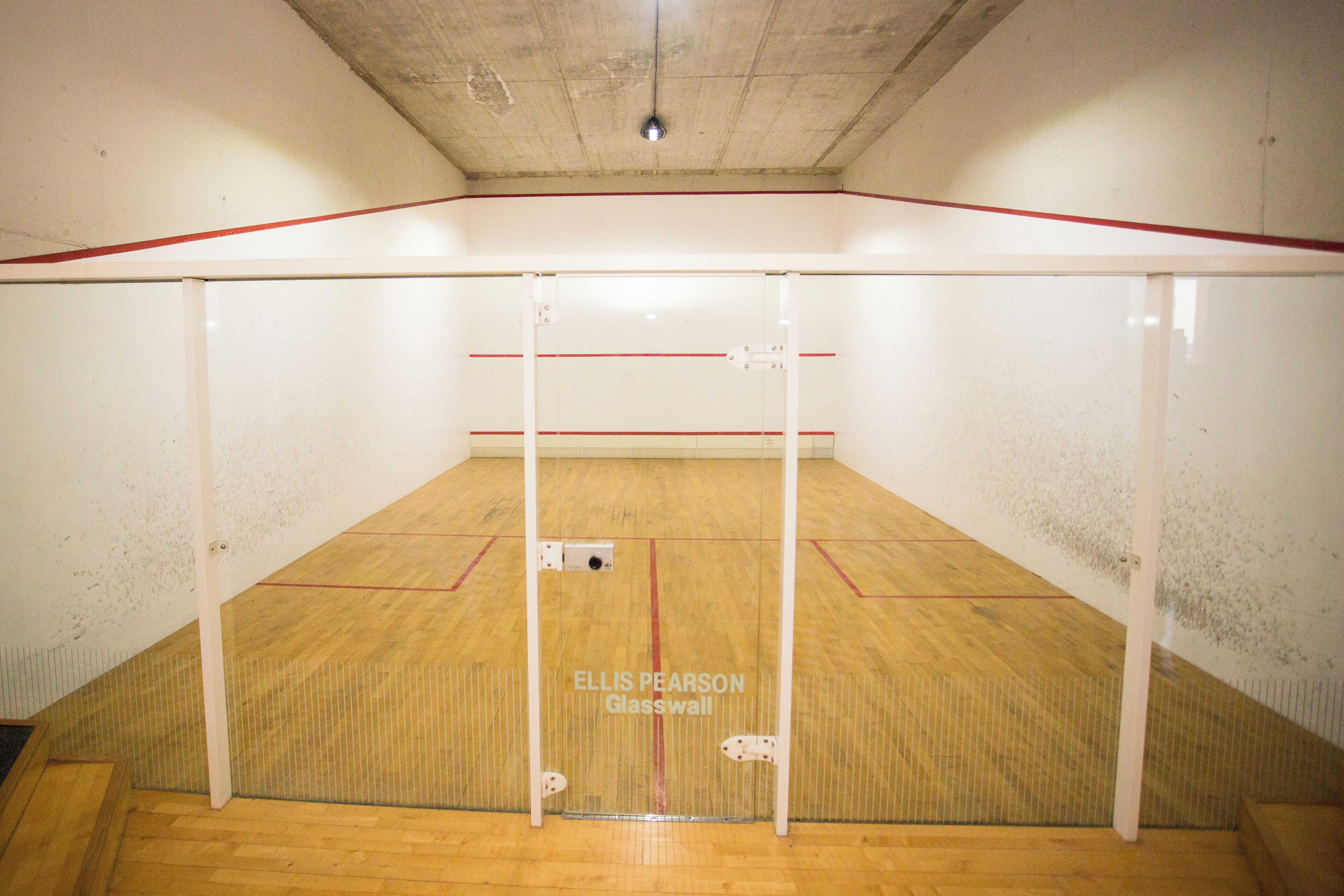 Una cancha de squash con piso de madera y paredes de vidrio, identificada como "ELLIS PEARSON Glasswall".