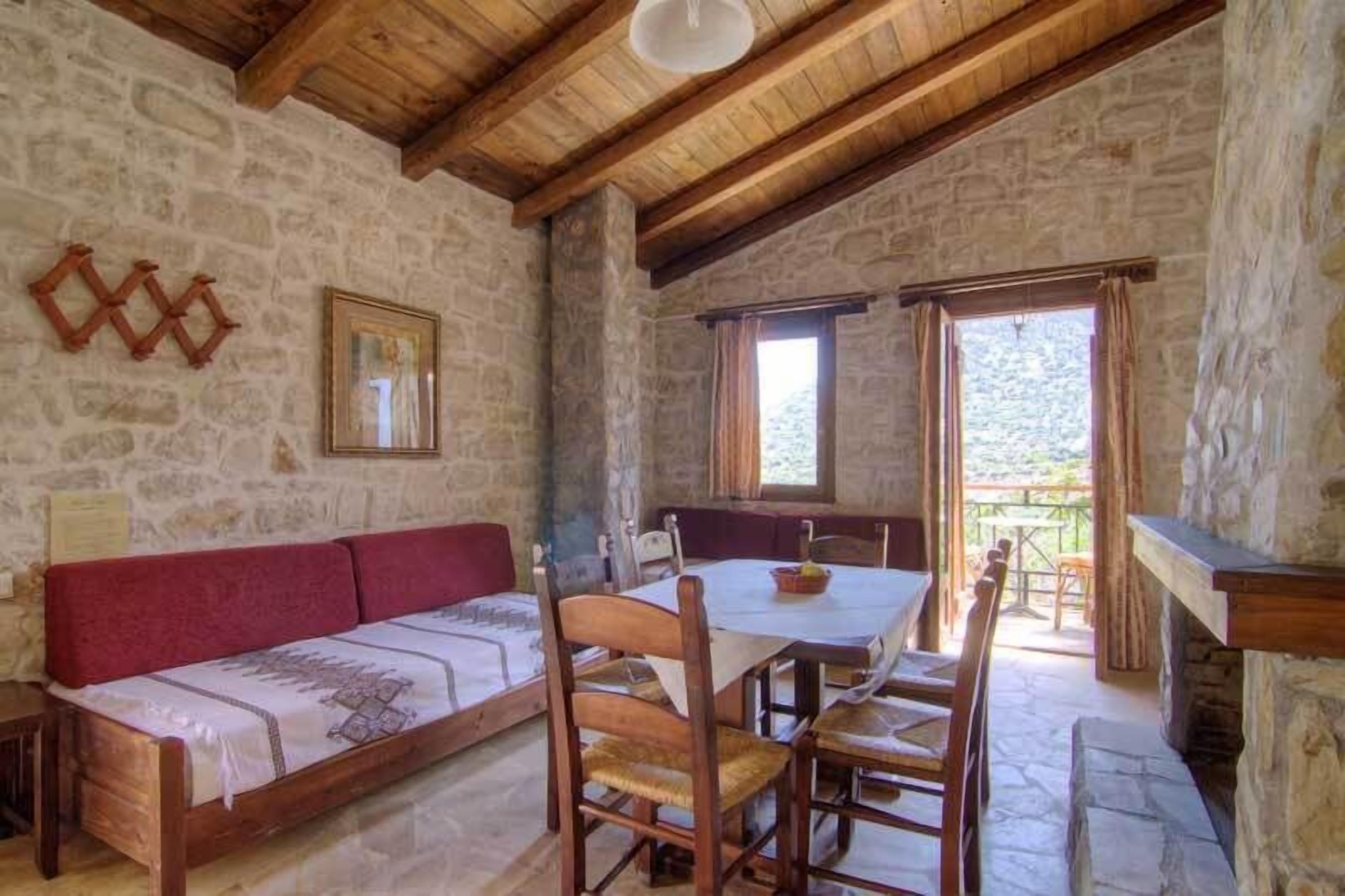 Ein rustikaler Steinraum mit Holzboden, einem roten Sofa, einem Esstisch mit Stühlen und Blick durch ein Fenster.