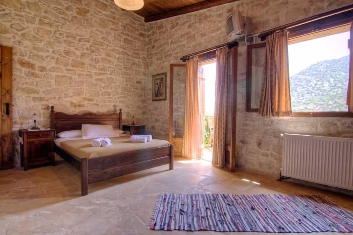A stone-walled bedroom with a wooden bed, a radiator, and a window with curtains.