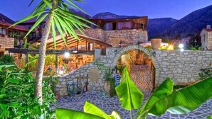 A stone archway labeled "Stone Village" leading to a building with a red roof and stone walls, surrounded by lush greenery and a mountainous backdrop. - Stone Village (Mylopotamos)