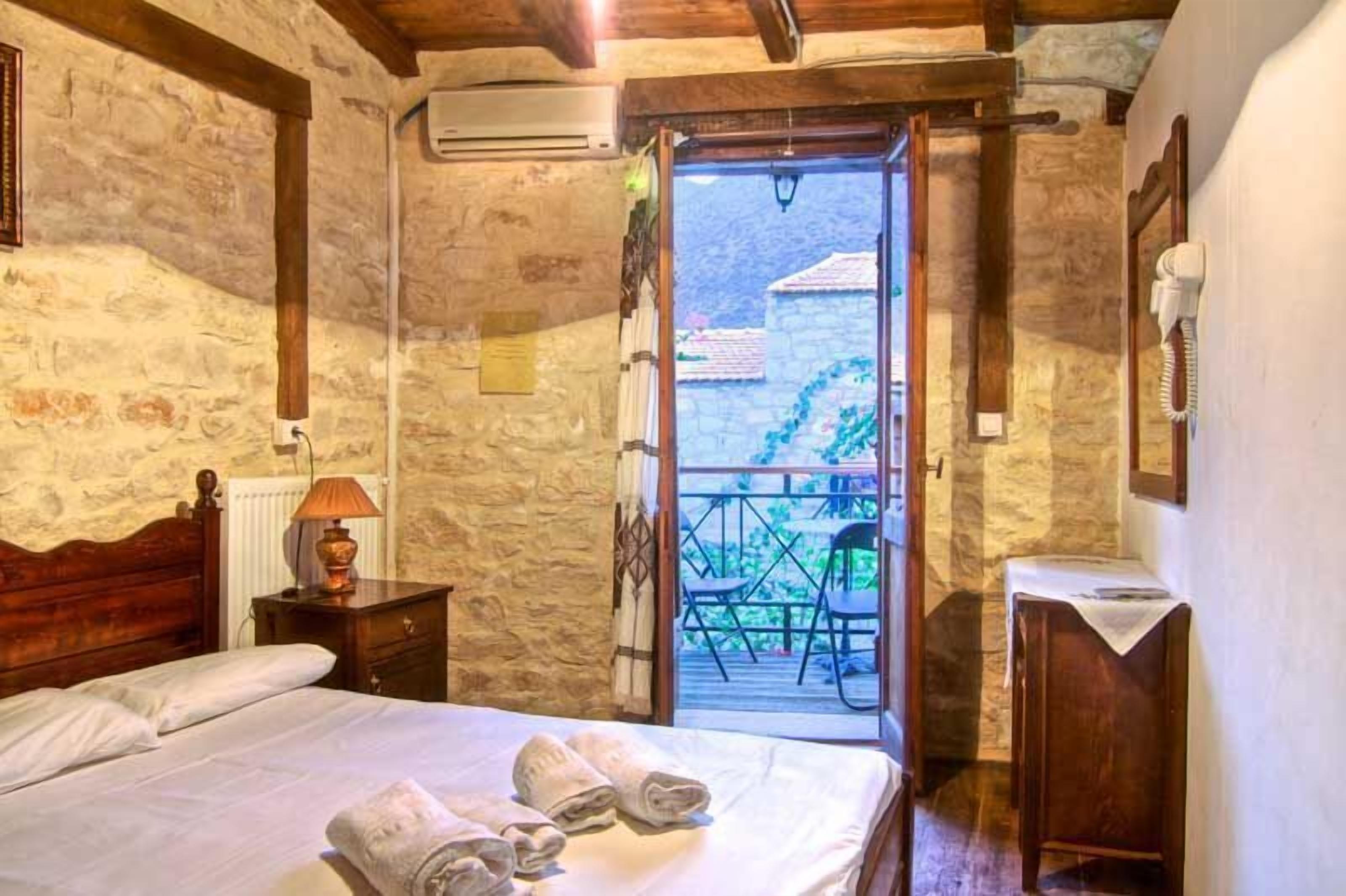 A bedroom with a stone wall, wooden bed, bedside lamp, and a view of a balcony with chairs.