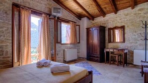A room with a bed, wooden desk, stone walls, and a view of the outdoors. - Stone Village (Mylopotamos)