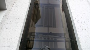Una estatua de piedra de un niño sosteniendo una esfera, ubicada en un recinto de vidrio reflectante en el interior de un edificio.