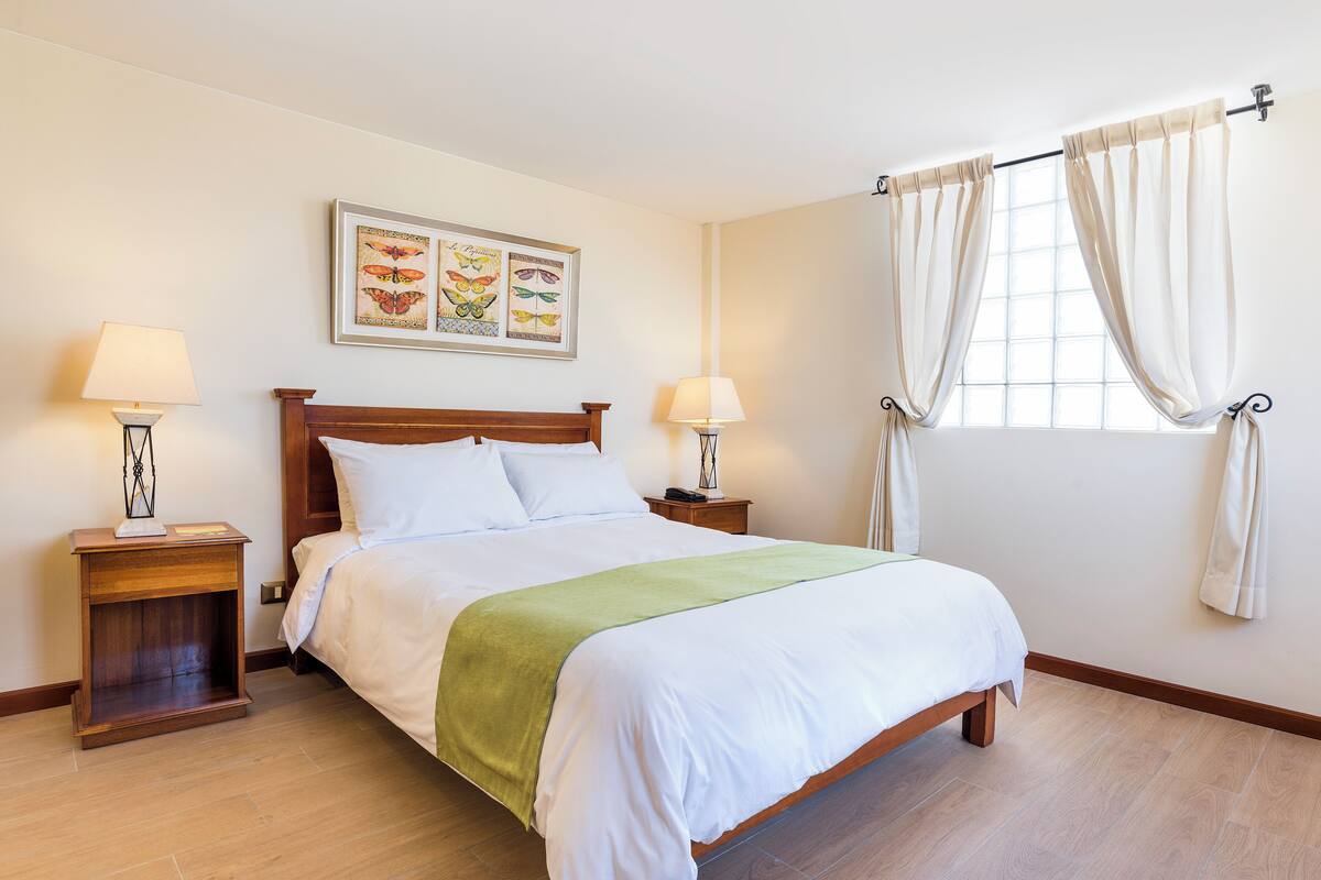 Una cama bien tendida con un plaid verde, dos mesitas de noche con lámparas y una ventana con cortinas.