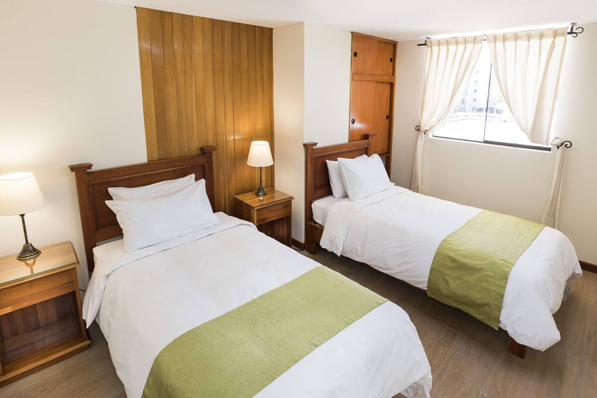 Habitación de hotel con dos camas individuales, cabeceros de madera, una mesita de noche con lámpara, un armario de madera y una ventana con cortinas.