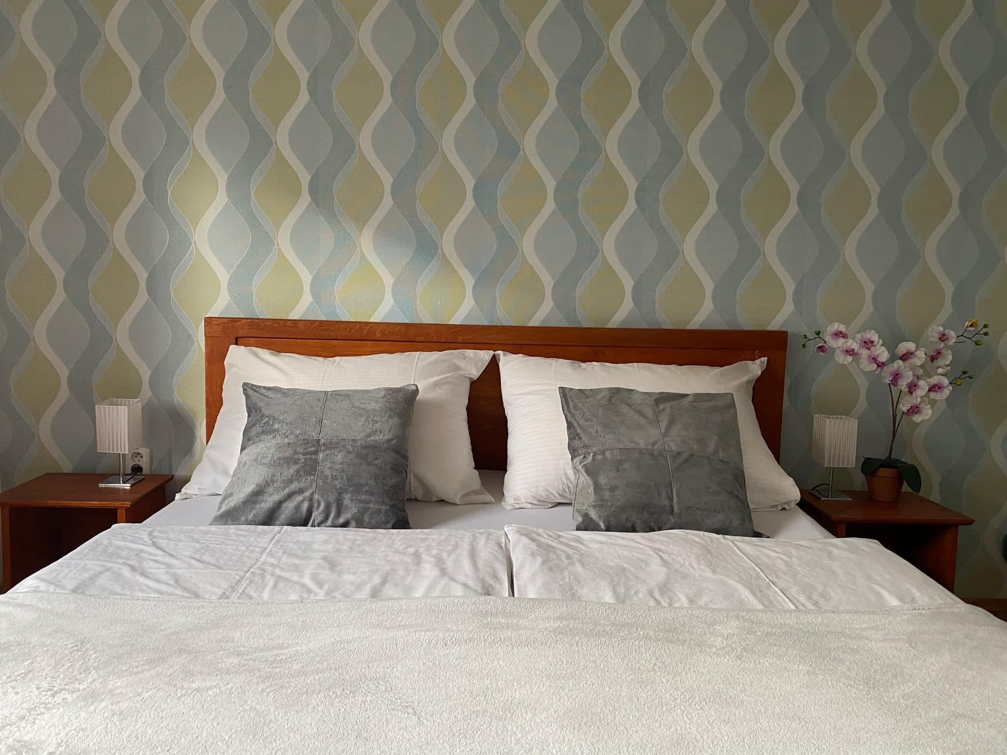 A neatly made bed with a wooden headboard, two bedside tables with lamps, and a patterned wallpaper.