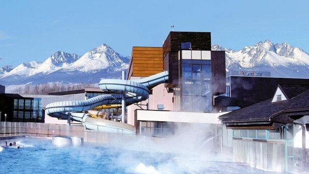 A modern hotel with a hot tub area and water slides, set against a backdrop of snow-capped mountains. - Penzion Plesnivec (Poprad)